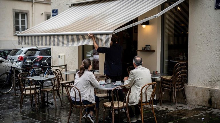 Dvoje gostiju sedi u bašti kafića dok konobar spušta prugastu tendu