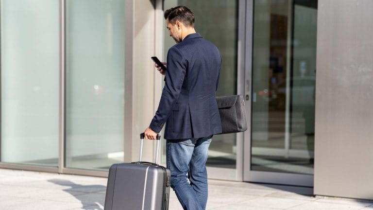 Muškarac u tamnom sakou vuče kofer i gleda u mobilni ispred staklene poslovne zgrade