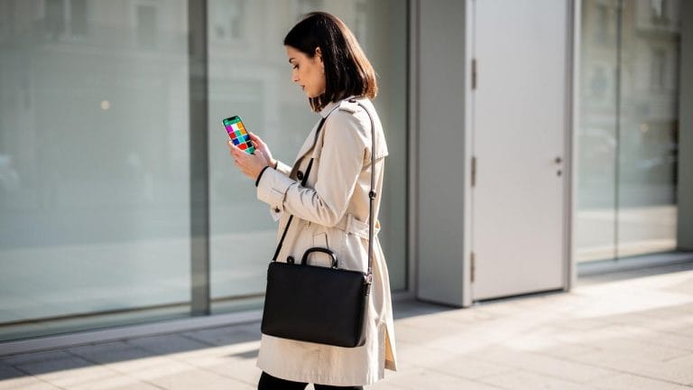 Žena u bež baloneru stoji i gleda u mobilni telefon držeći crnu poslovnu torbu
